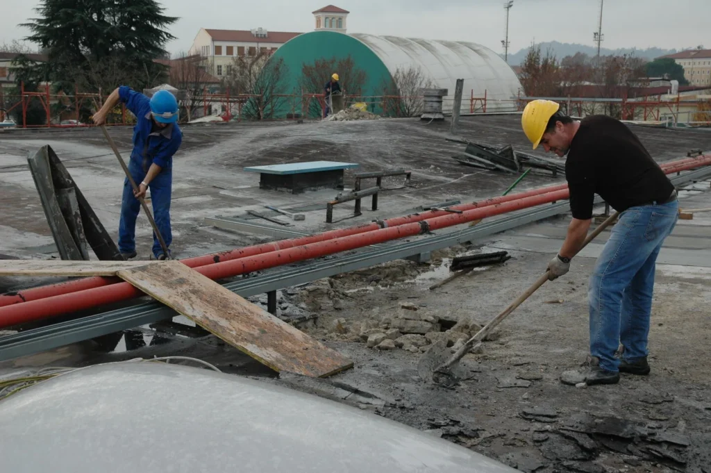 Two construction workers in hard hats remove debris on a flat rooftop, one with a shovel and the other with a pry bar, amid red pipes and rubble.